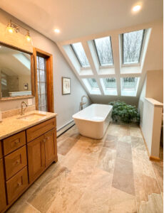 Bathroom remodel with light tile floors, woodgrain vanity with light stone countertop, soaking tub along far wall, under slanted ceiling with two rows of skylights.